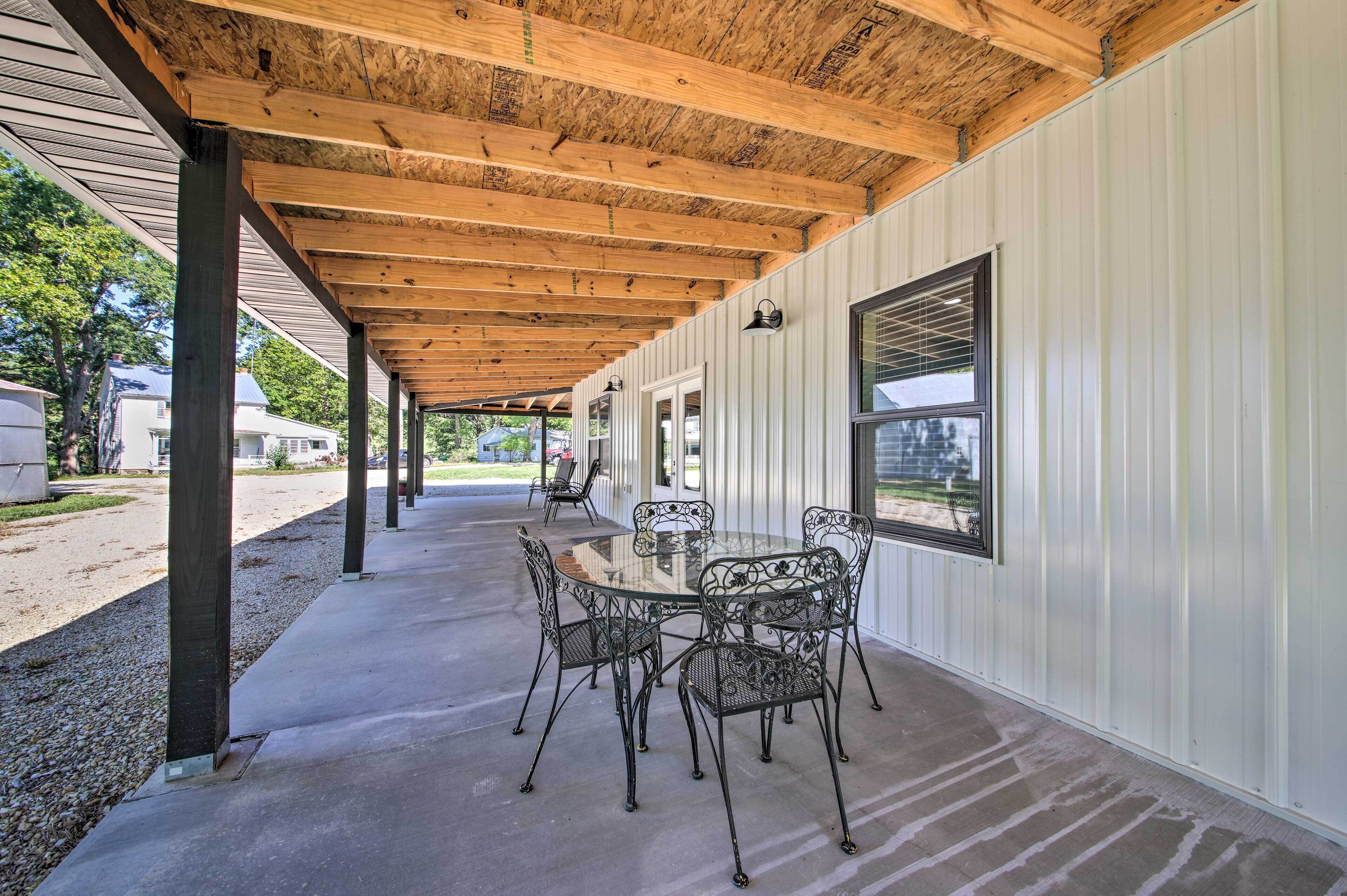Un patio coperto con tavolo in vetro e sedie in metallo, travi in legno a soffitto e vista su una strada residenziale.