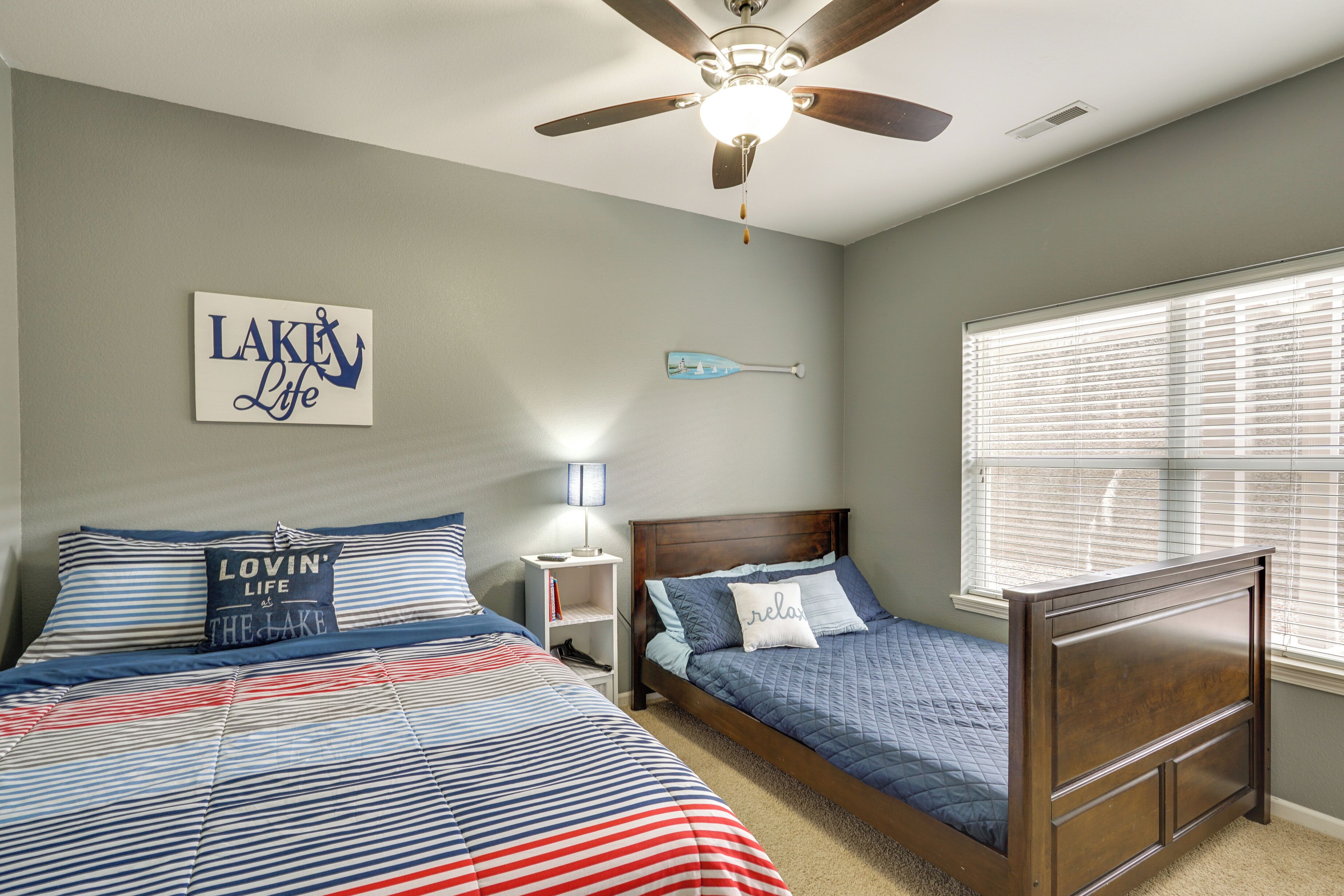 A bedroom with two beds, a ceiling fan, a nightstand, a wall-mounted paddleboard, and a window with blinds.