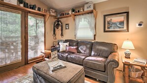 A cozy living room with a leather sofa, a coffee table, and a lamp. There are shelves with books and decorative items, and a window with blinds.