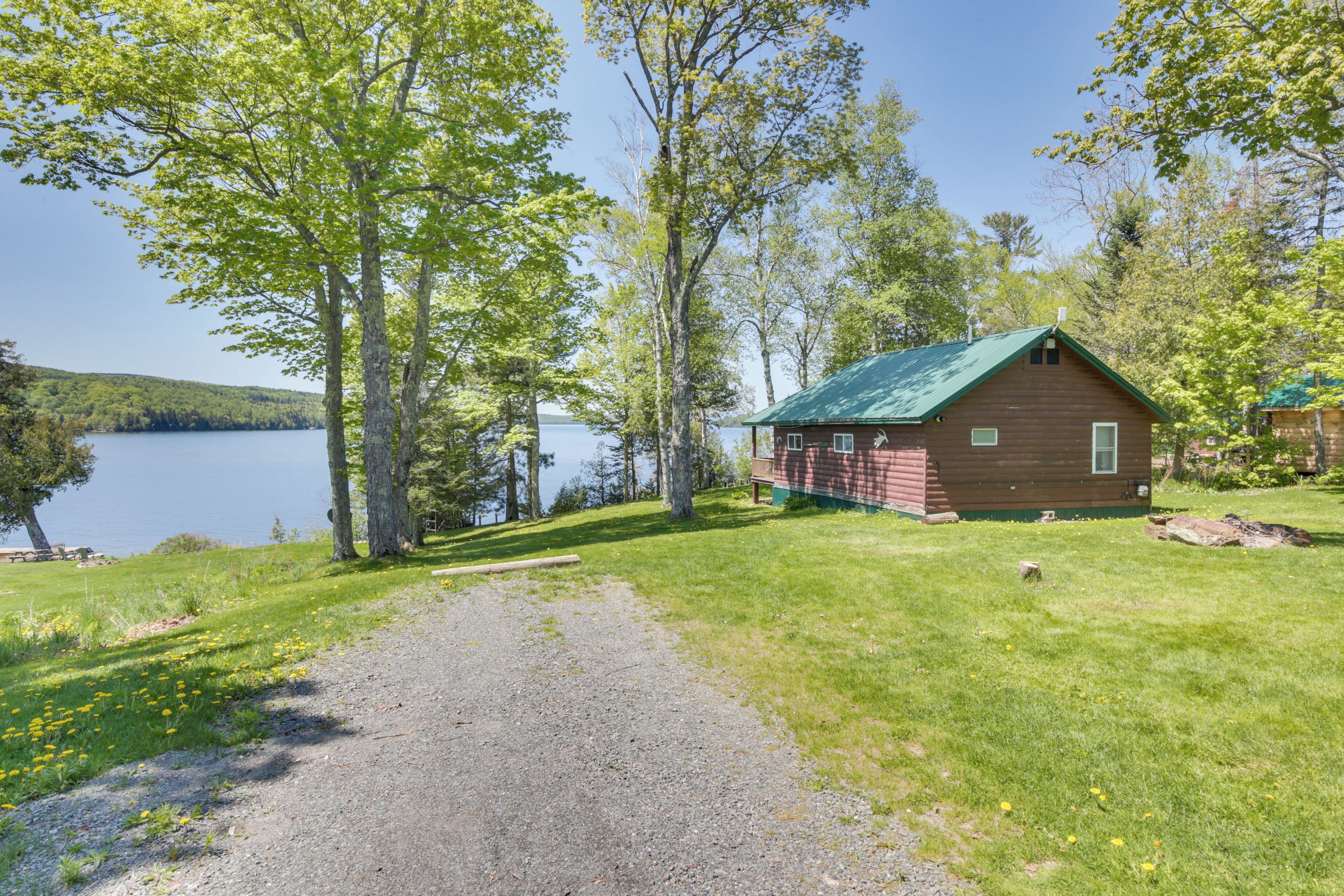 A cabin by a lake with a gravel path leading to it, surrounded by trees and grass.