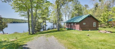 A cabin by a lake with a gravel path leading to it, surrounded by trees and grass.