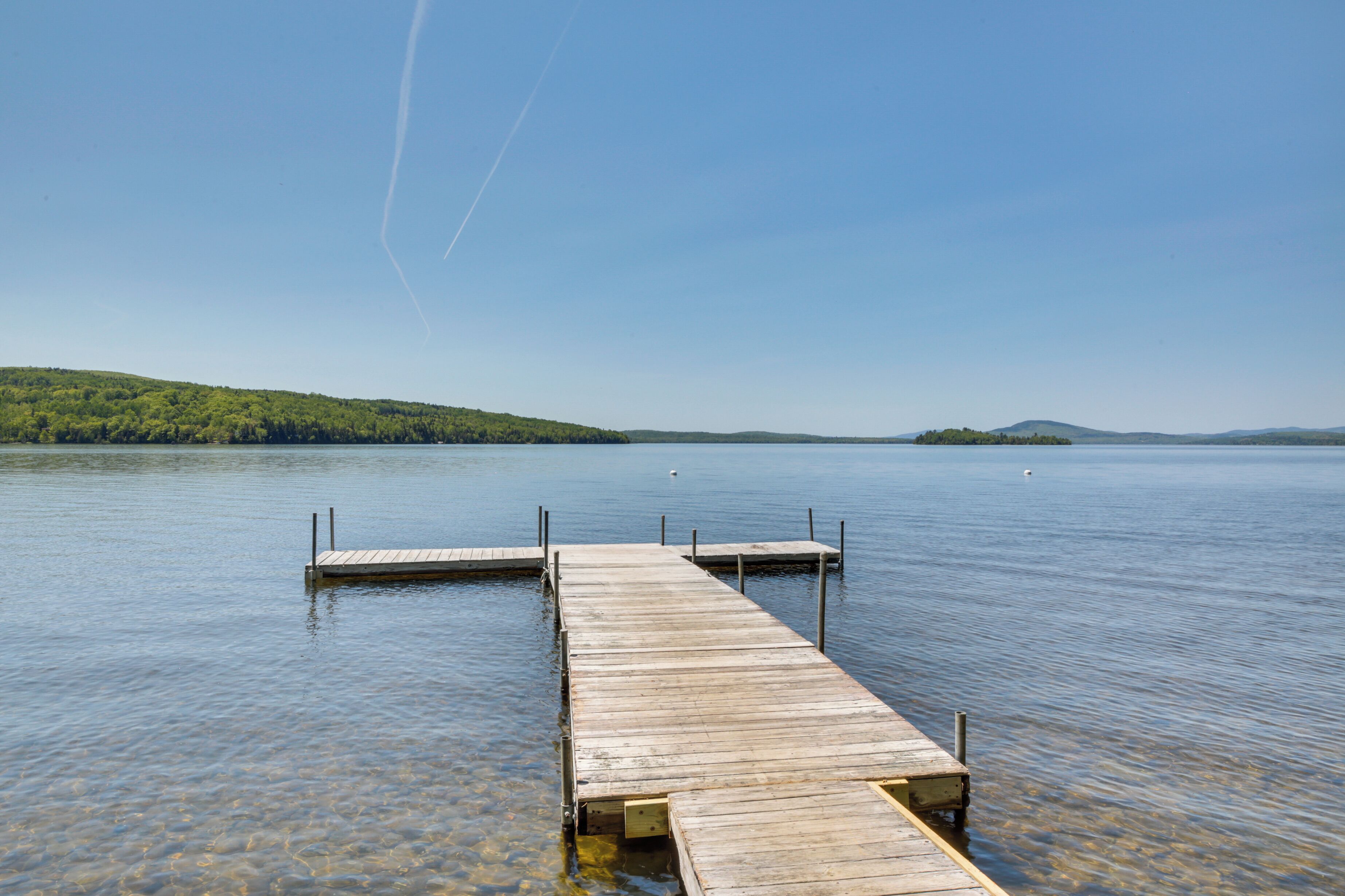 A wooden dock extending into a calm lake with clear water, surrounded by green hills.