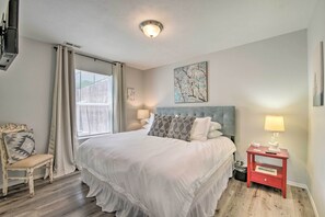 A bedroom with a large bed, a nightstand with a lamp, a chair, and a window with curtains.