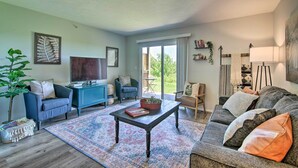 Een woonkamer met een bank, fauteuil, salontafel en een televisie. Een groot raam zorgt voor daglicht en uitzicht op het buiten.