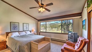 A bedroom with a bed, a wicker side table, a television, a chair, and a large window with a view of trees and mountains. - Quiet Unit w/ Golf Course & Mtn Views in Graeagle! (Blairsden-Graeagle)