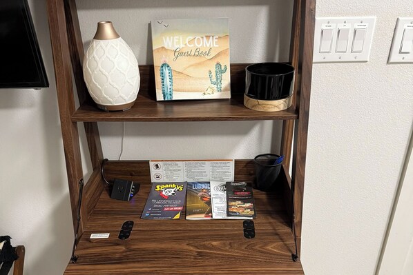 A wooden shelf with a welcome sign, a vase, and a candle. Below, there are brochures and a remote control.