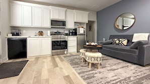 A modern kitchen with white cabinets, a microwave, oven, and coffee maker. There is a gray sofa with patterned cushions and a round mirror on the wall. - Relaxing Lubbock Studio ~ 6 Mi to Texas Tech (Lubbock)