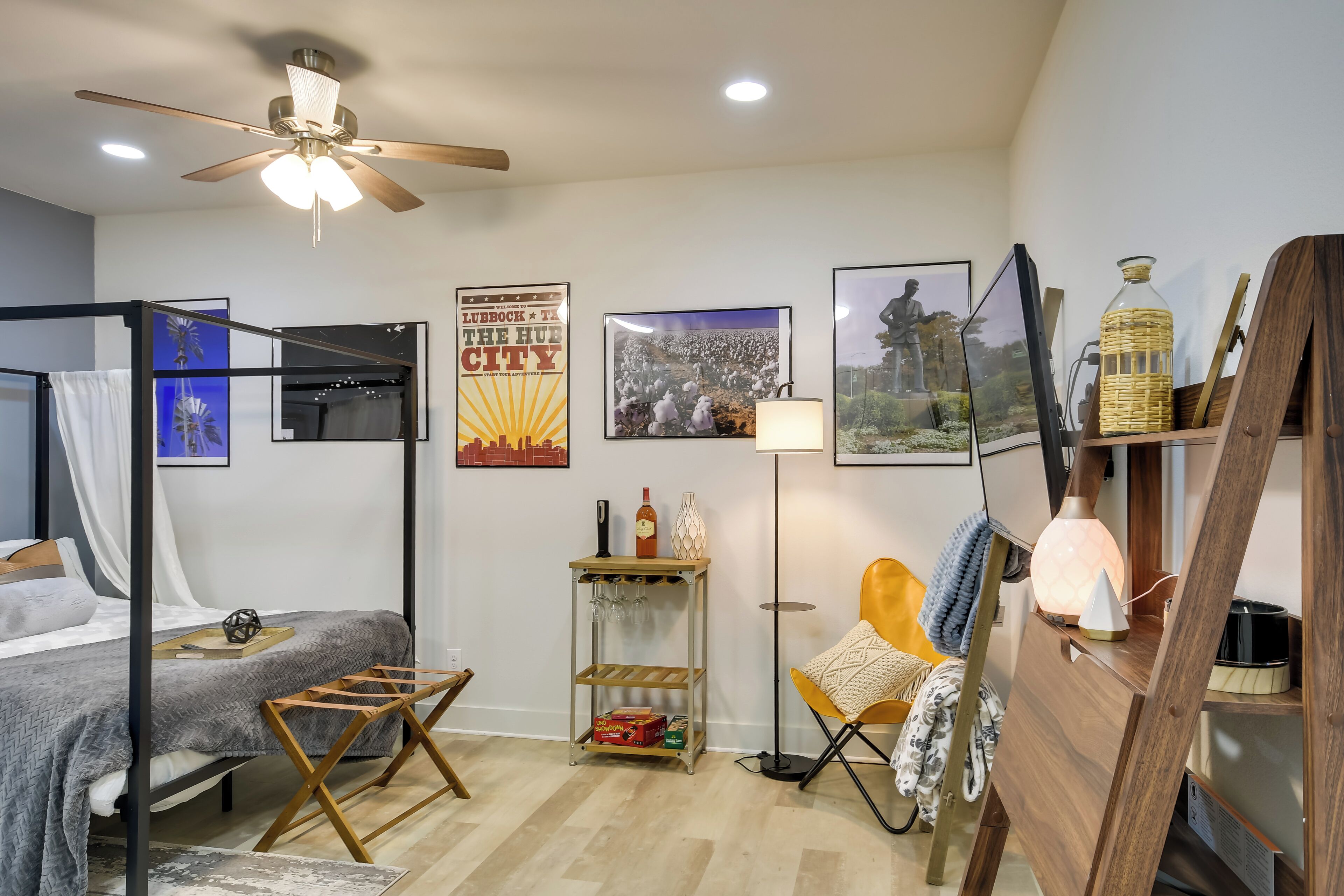 A modern bedroom with a bed, a standing fan, a small table with a lamp, and a poster on the wall.