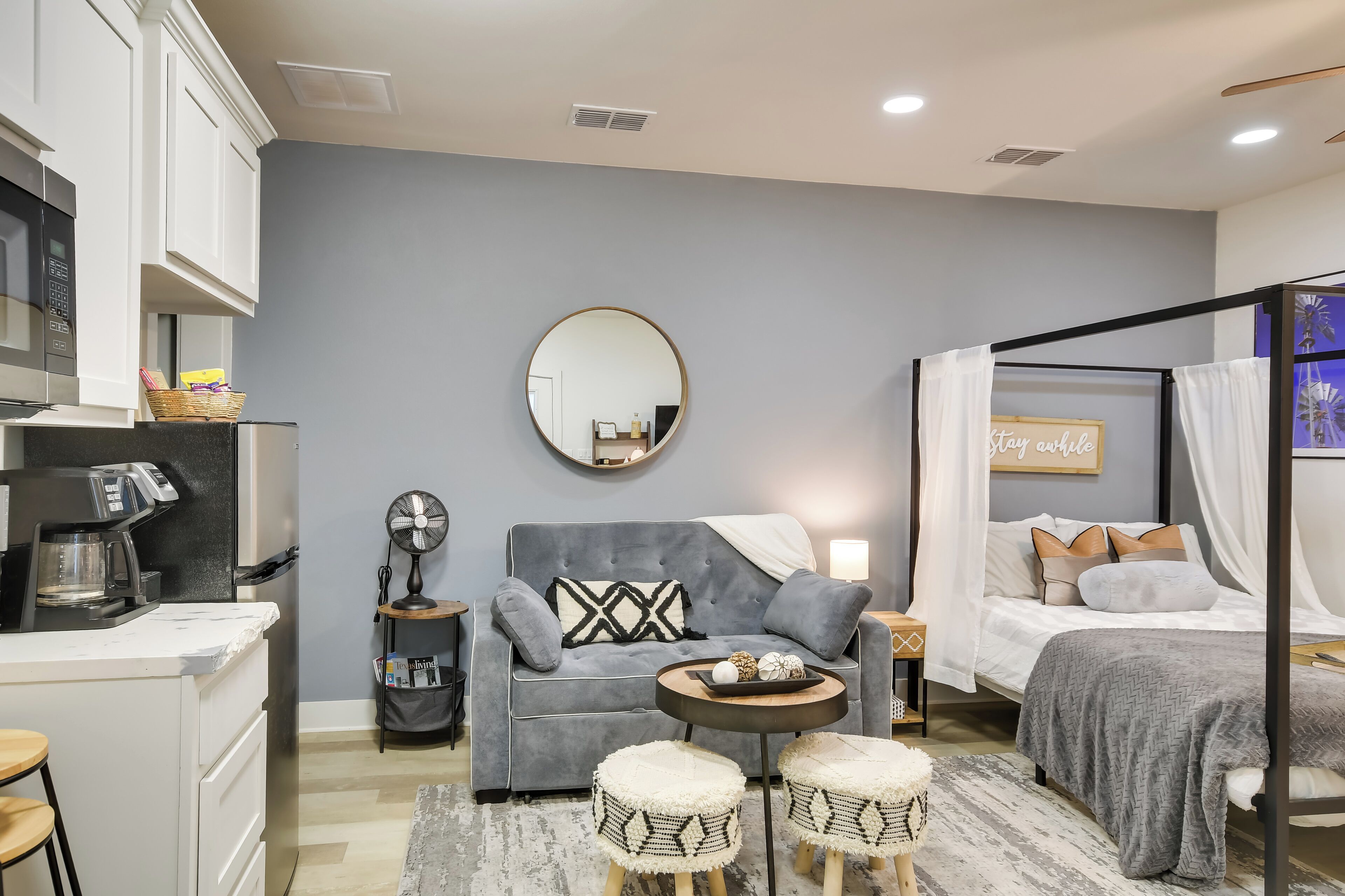 A compact bedroom with a grey sofa, a round mirror, a bed with a canopy, and a small round table with decorative items.