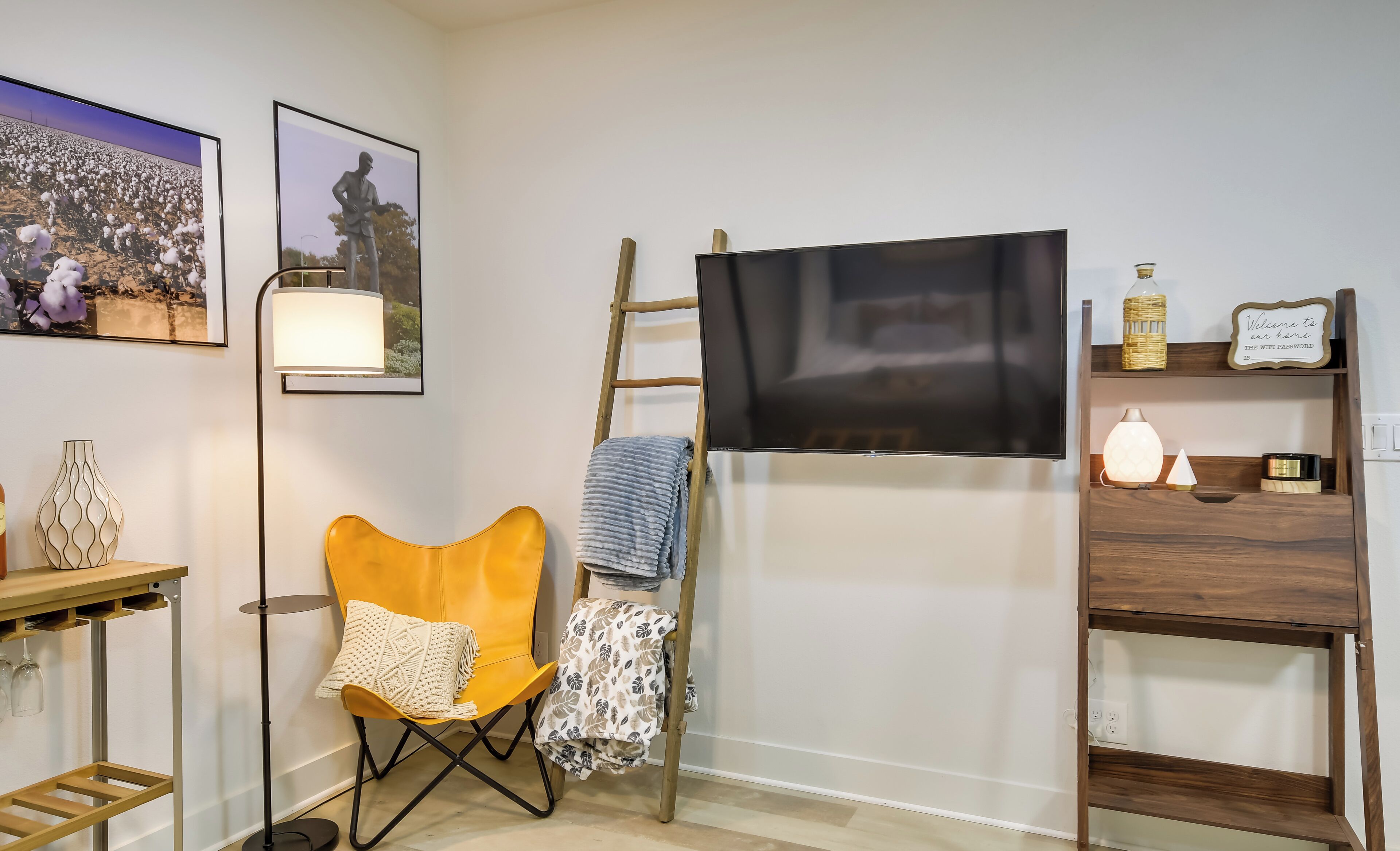 A room with a mounted TV, a yellow chair, a ladder, a shelf with decorative items, and framed pictures on the wall.