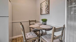 A corner dining area with a wooden table and four chairs, each with a cushioned seat. A small potted plant is on the table, and a framed artwork hangs on the wall above.