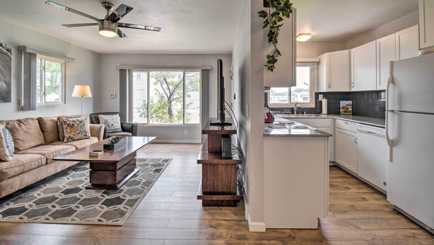 A modern living room with a sofa, coffee table, and a TV. There is a kitchen area with white cabinets and a refrigerator.