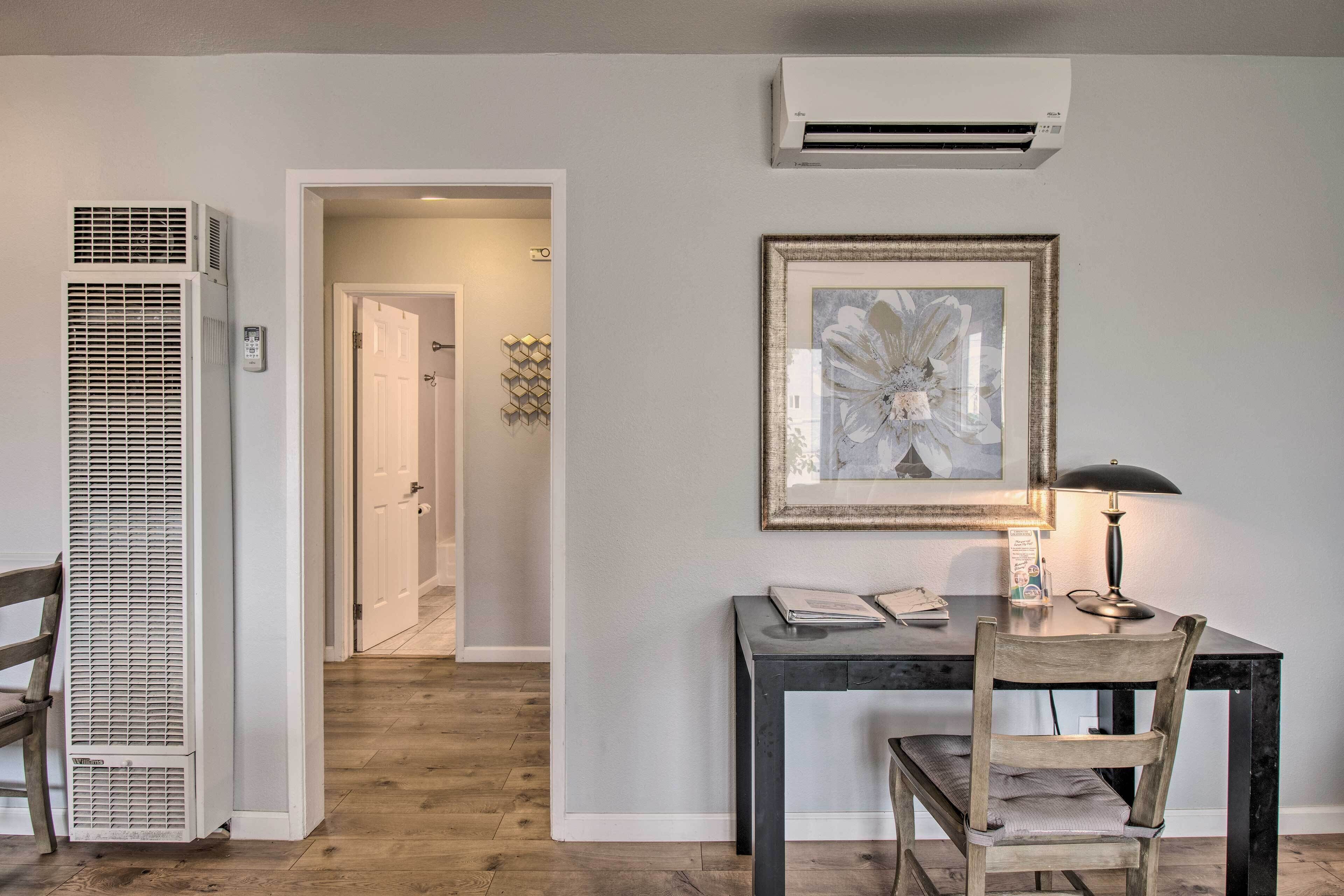 A room with a desk, chair, lamp, air conditioning unit, and a hallway leading to another room.