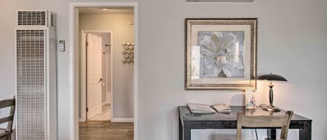 A room with a desk, chair, lamp, air conditioning unit, and a hallway leading to another room.