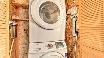 Lavadora compacta de dos puertas con panel de control y una etiqueta que indica 10 años de garantía, instalada en un cuarto de servicio de madera con un estante y un trapeador.