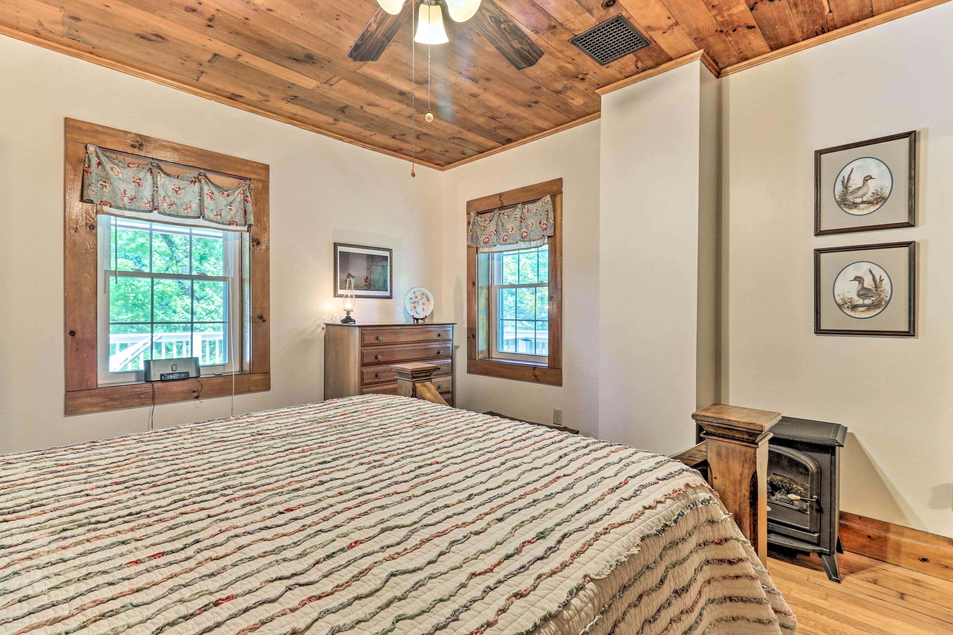 A bedroom with a large bed, two windows with curtains, a dresser, a desk, and a wall-mounted heater.
