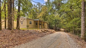 A single-story cabin with a gravel driveway, surrounded by dense forest. - Quaint Tallassee Cabin: Stone Creek On-site! (Tallassee)