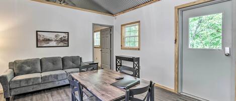 A room with a wooden dining table, black chairs, a gray sofa, a framed picture on the wall, and a door with a window.