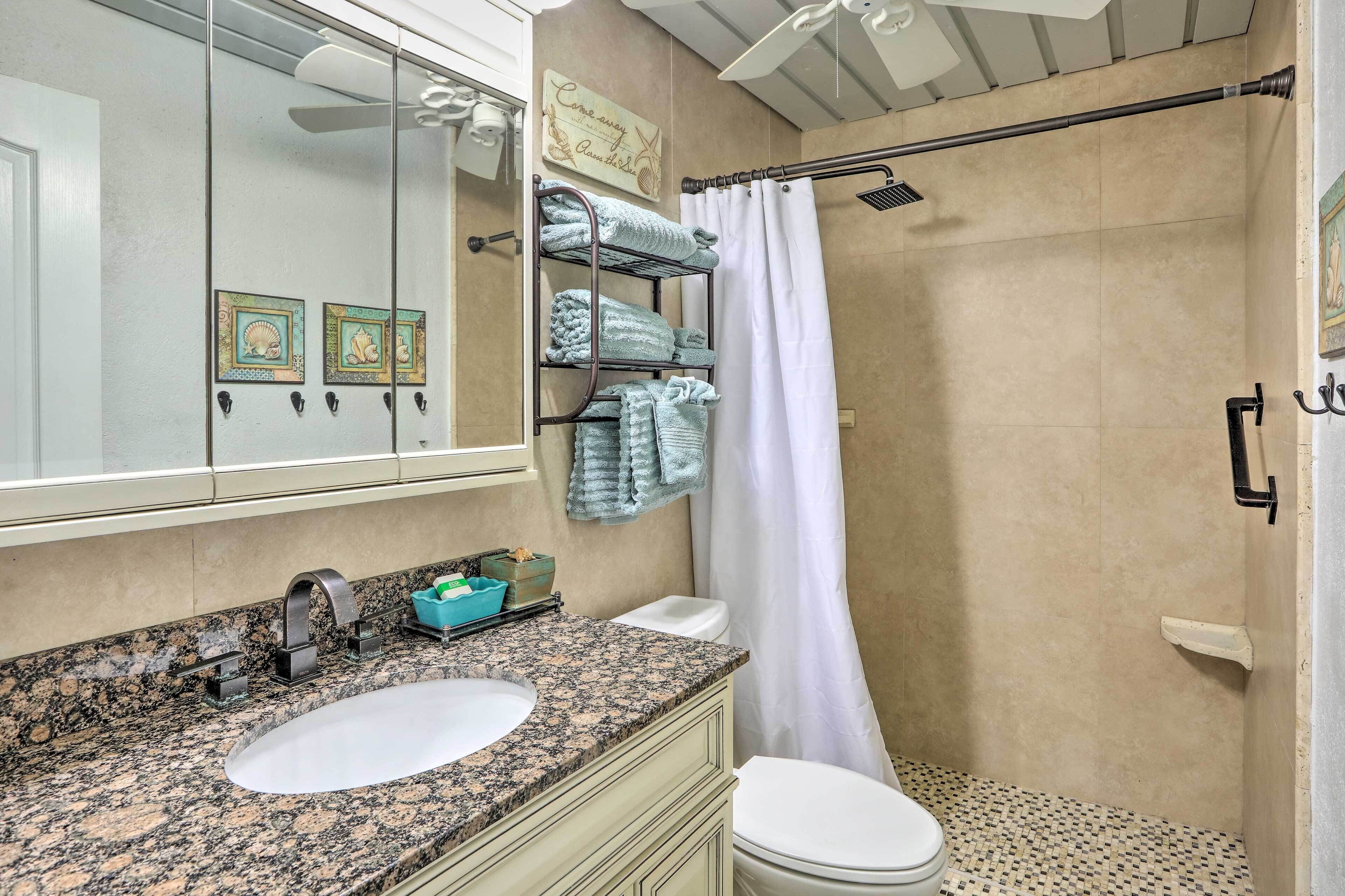 A bathroom with a granite countertop sink, a white toilet, a glass shower enclosure, and a towel rack with folded towels.