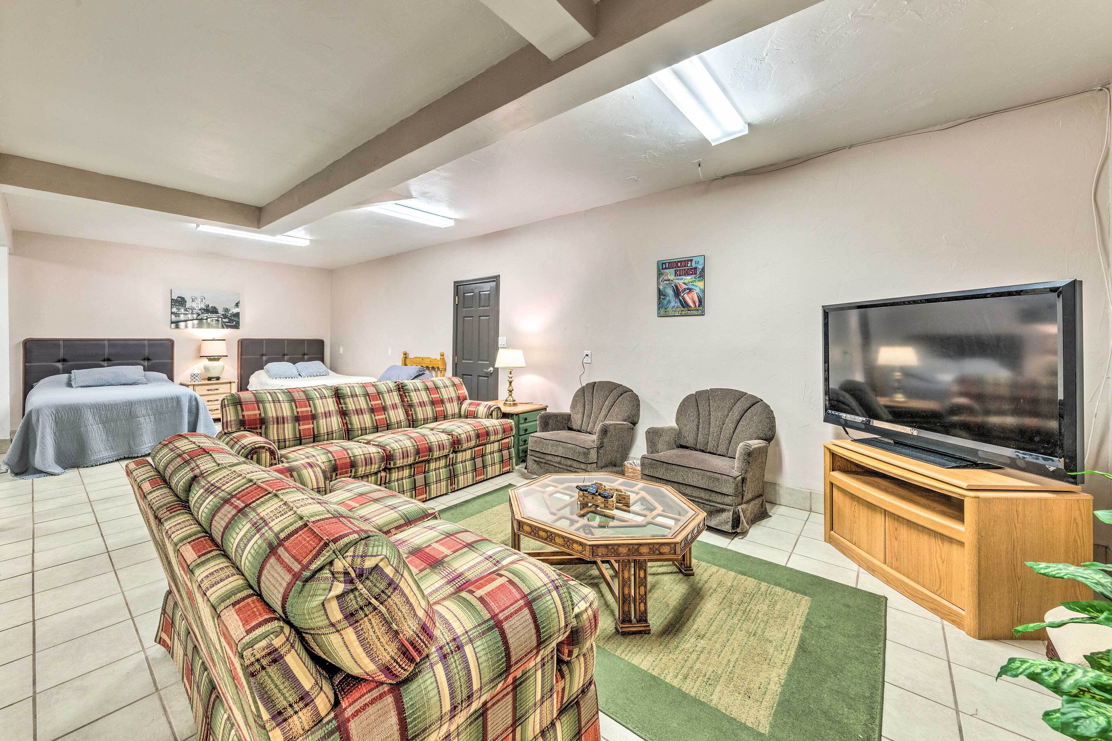 A living room with a sofa set, armchairs, a coffee table, a TV on a wooden stand, and a bed with a blue cover.