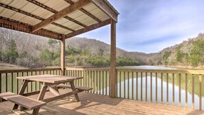 A wooden deck with a picnic table and a metal roof structure overlooking a calm lake with forested hills in the background.