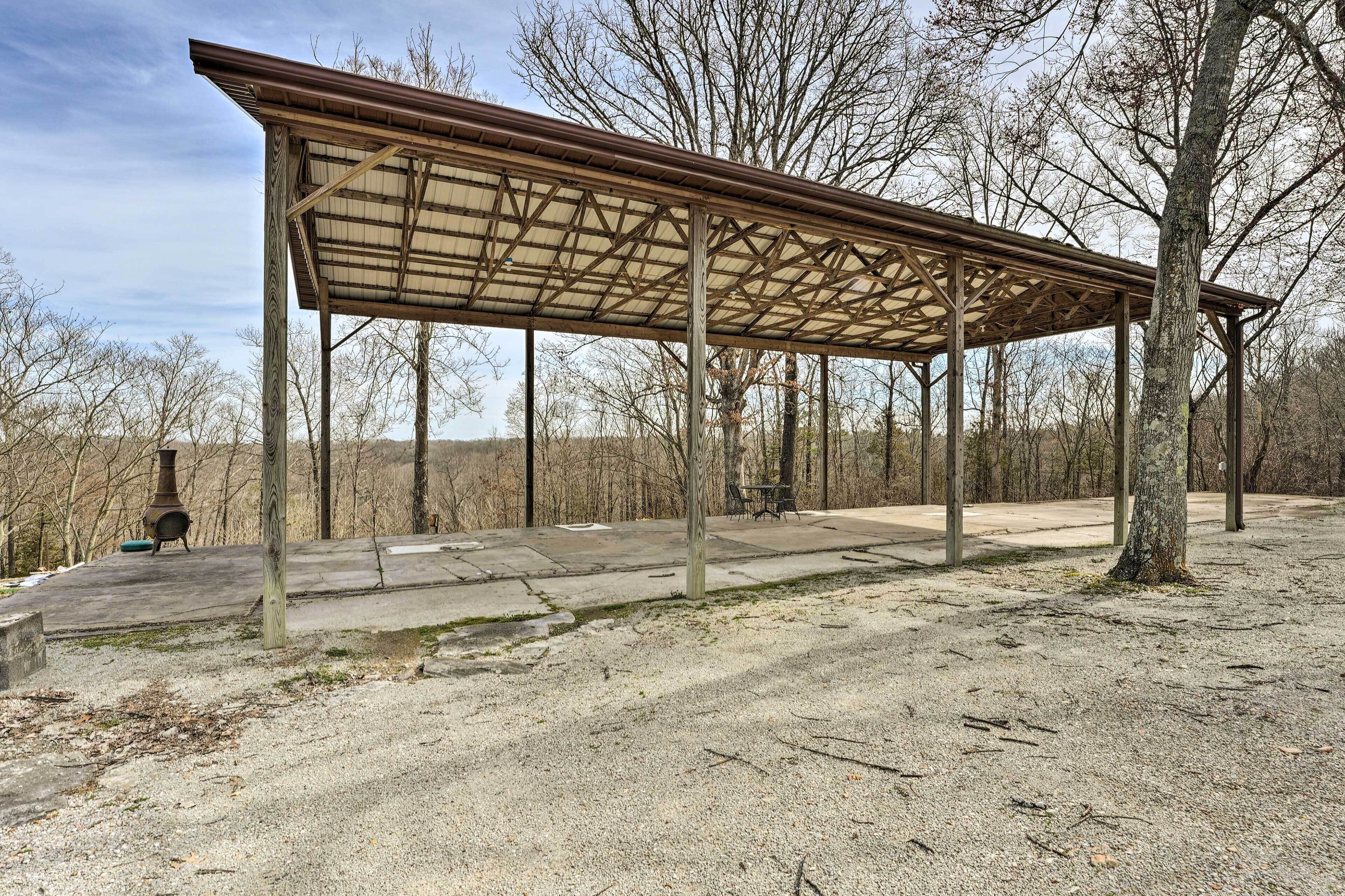 A covered picnic area with a metal roof and concrete floor, surrounded by trees.