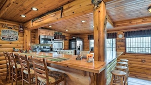 A log cabin interior with a central wooden bar, bar stools, a kitchen area with modern appliances, and a large window with a view. - Rustic Cabin in Roaring River State Park! (Eagle Rock)