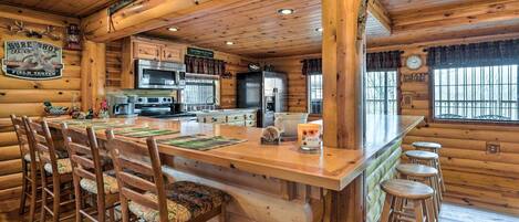 Interior de una cabaña de troncos con una barra de madera central, taburetes de bar, una zona de cocina con electrodomésticos modernos y un ventanal con vistas.
