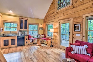 A cozy cabin interior with wooden walls, a kitchen with a dishwasher, a dining area with a table and chairs, and a red sofa with a striped pillow. - Romantic Asheville Area Cabin w/ Deck & Hot Tub! (Leicester)