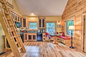 A cabin interior with wooden floors, a kitchen with a stove and dishwasher, a dining area with a table and chairs, and a window with a view of trees. - Romantic Asheville Area Cabin w/ Deck & Hot Tub! (Leicester)