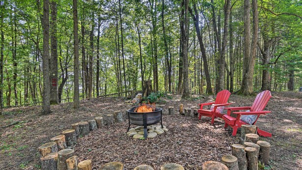 Waldlandschaft mit Lagerfeuerstelle, umgeben von Baumstämmen, und zwei roten Adirondack-Stühlen.