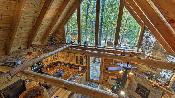 A rustic cabin interior with a high ceiling, wooden beams, a stone fireplace, and a view of the forest through large windows.