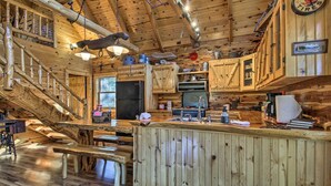 A rustic cabin interior with a wooden staircase, a bar area, and a kitchen with wooden cabinets and a large clock.