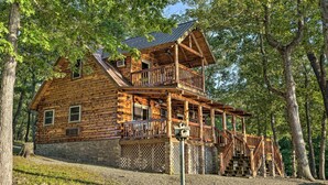 A log cabin with a balcony and a stone foundation, surrounded by trees.