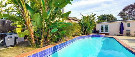 Uma piscina com azulejos azuis e laranja, rodeada por bananeiras e uma casa.