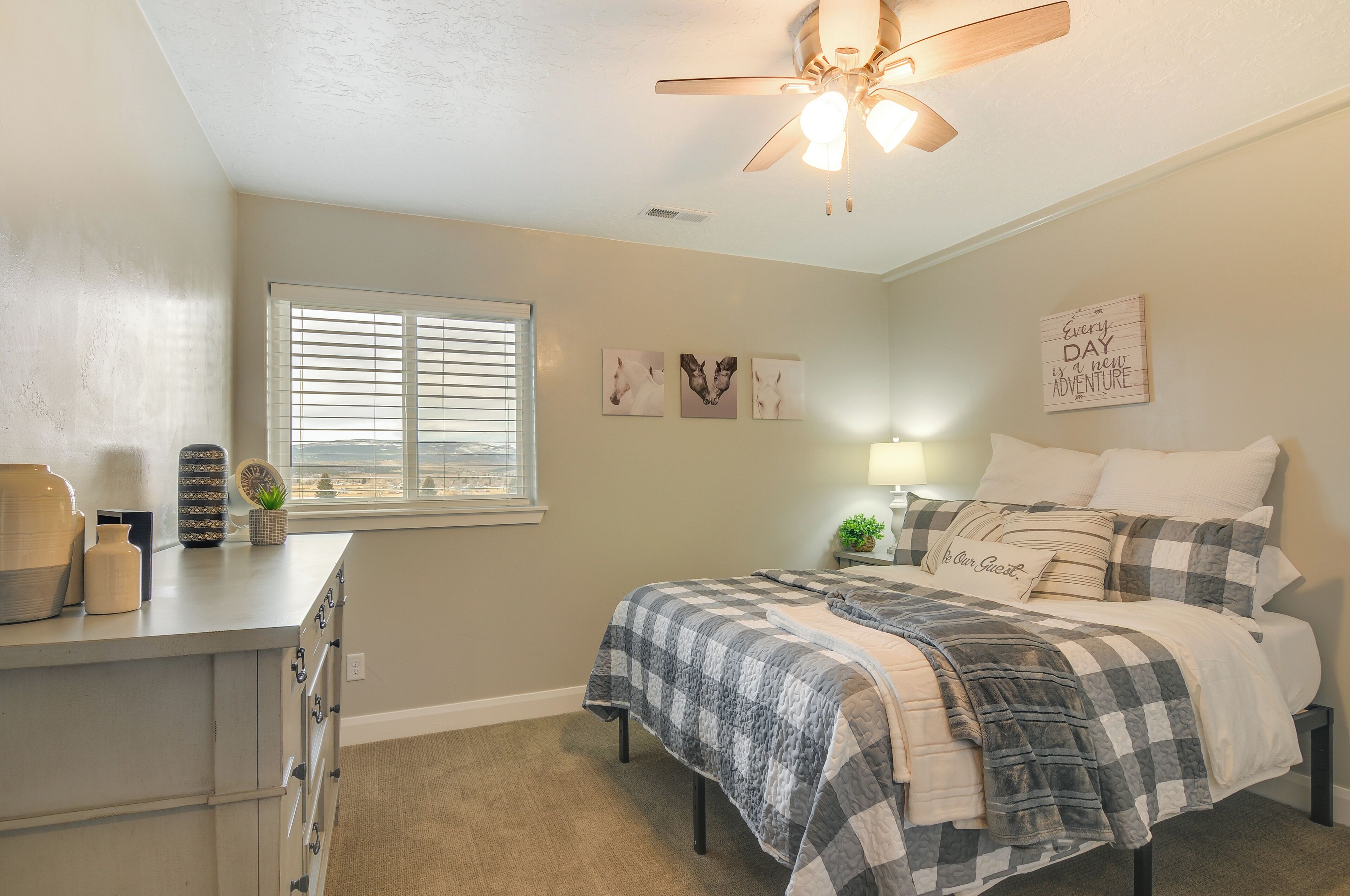 A bedroom with a bed, a ceiling fan, a window with blinds, a dresser, and wall art.