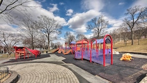 A playground with red and blue play structures, a paved area, and bare trees in a park. - Walk to Shops, Dining & City Park: Reading Apt (Reading)