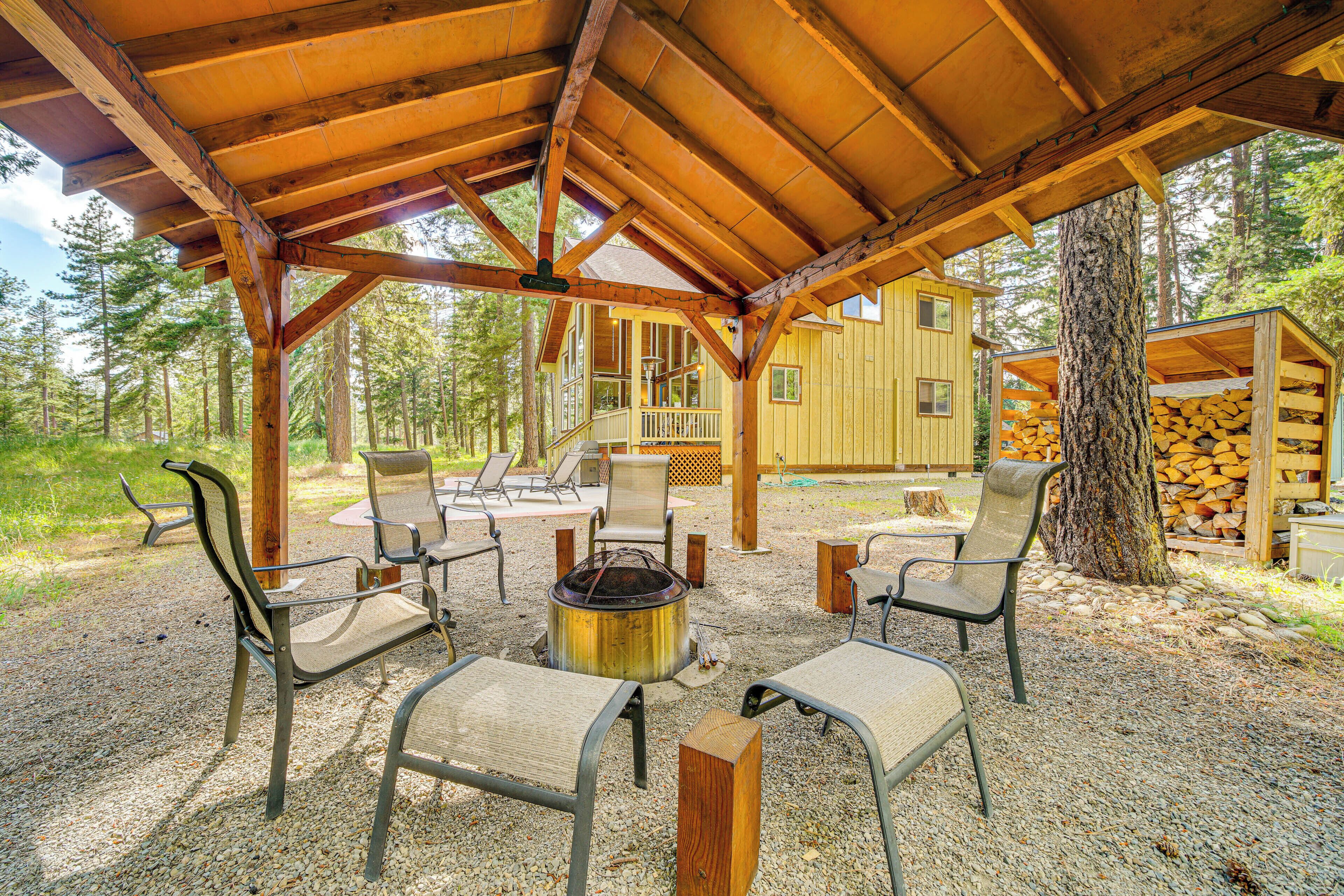 Un espace extérieur aménagé, abrité et doté de chaises et d’un foyer, entouré d’arbres et bordé d’un bâtiment en bois.