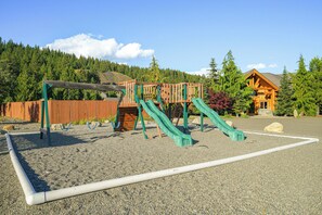 A playground with slides and a wooden structure, surrounded by a fence and a building with a sloped roof. - Ronald Vacation Rental Near Suncadia Resort (Cle Elum)