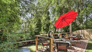 Eine Holzterrasse mit Gartenmöbeln und einem roten Sonnenschirm, umgeben von üppiger Vegetation und einem kleinen Teich.