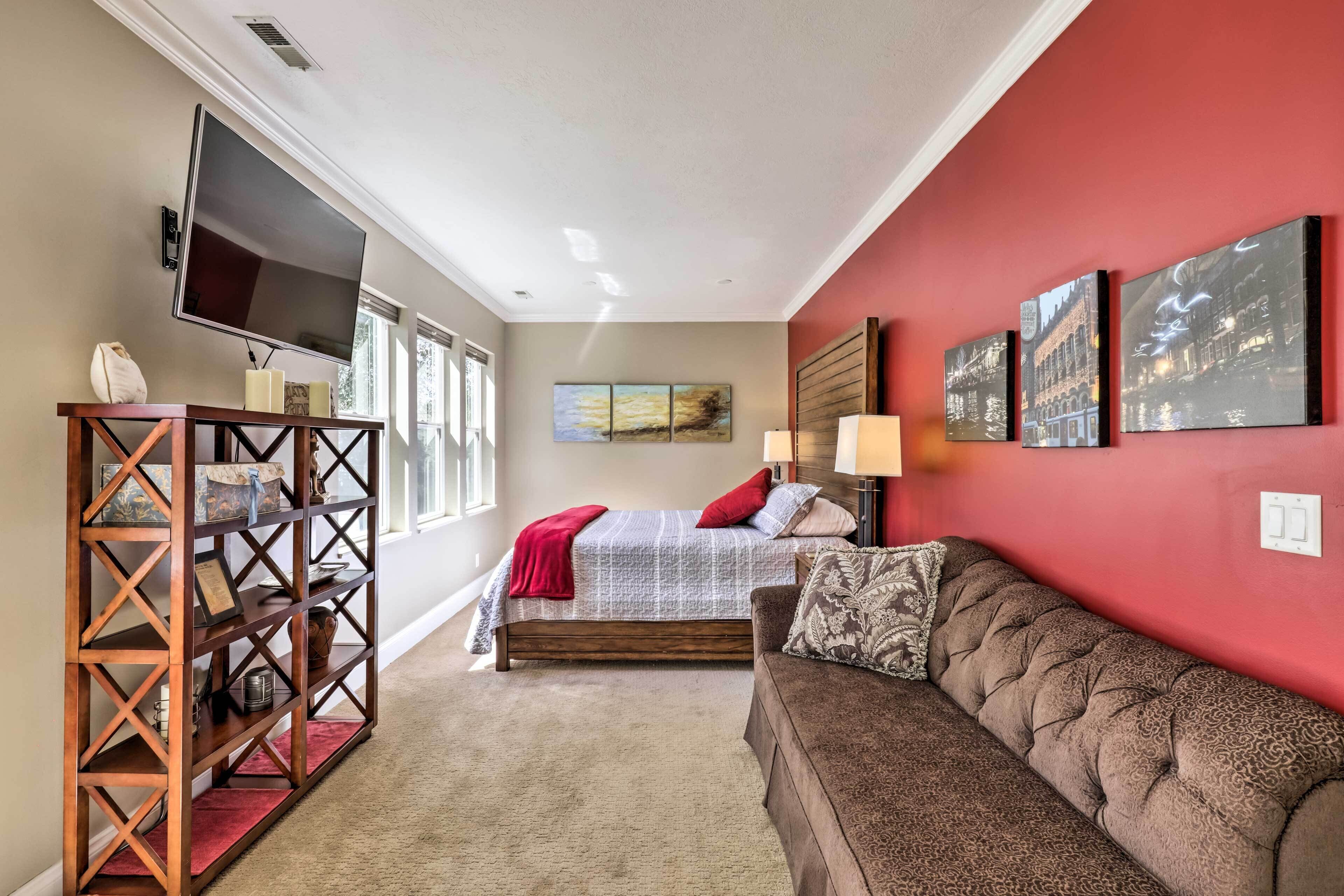 A bedroom with a bed, a red wall, a brown sofa, a TV, and artwork.