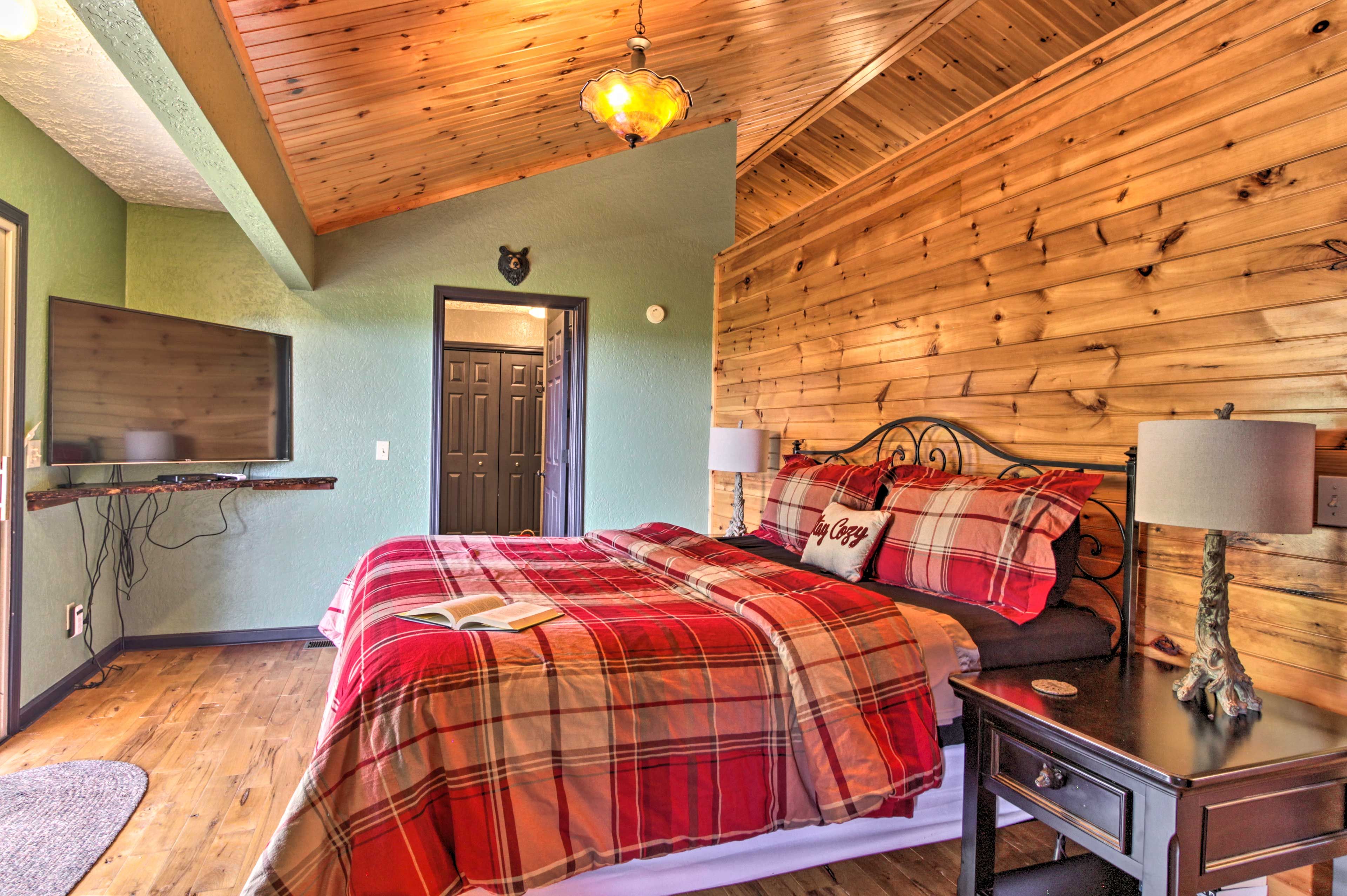 Una habitación acogedora con techo de madera, una cama con ropa de cama escocesa, una mesita de noche con una lámpara y un televisor montado en la pared.
