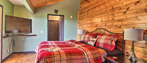 Una habitación acogedora con techo de madera, una cama con ropa de cama escocesa, una mesita de noche con una lámpara y un televisor montado en la pared.