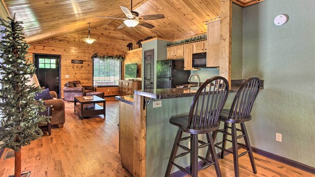A cozy cabin interior with a wooden ceiling, a small kitchen area, a dining space, and a living area with a Christmas tree.