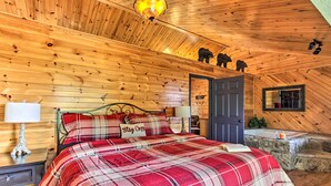 A cozy wooden cabin bedroom with a plaid bedspread, a nightstand with a lamp, a stone bathtub, and a door leading to another room.