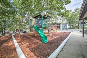 A playground with a slide and climbing structures, surrounded by trees and a paved walkway.