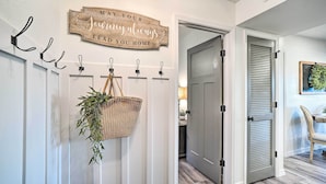 Un pasillo con un letrero de madera que dice “Que tu viaje siempre te lleve a casa”, una cesta tejida con follaje y una puerta con diseño persiana.