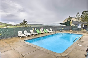 A swimming pool with lounge chairs and a shaded area, surrounded by a fence and a view of hills.