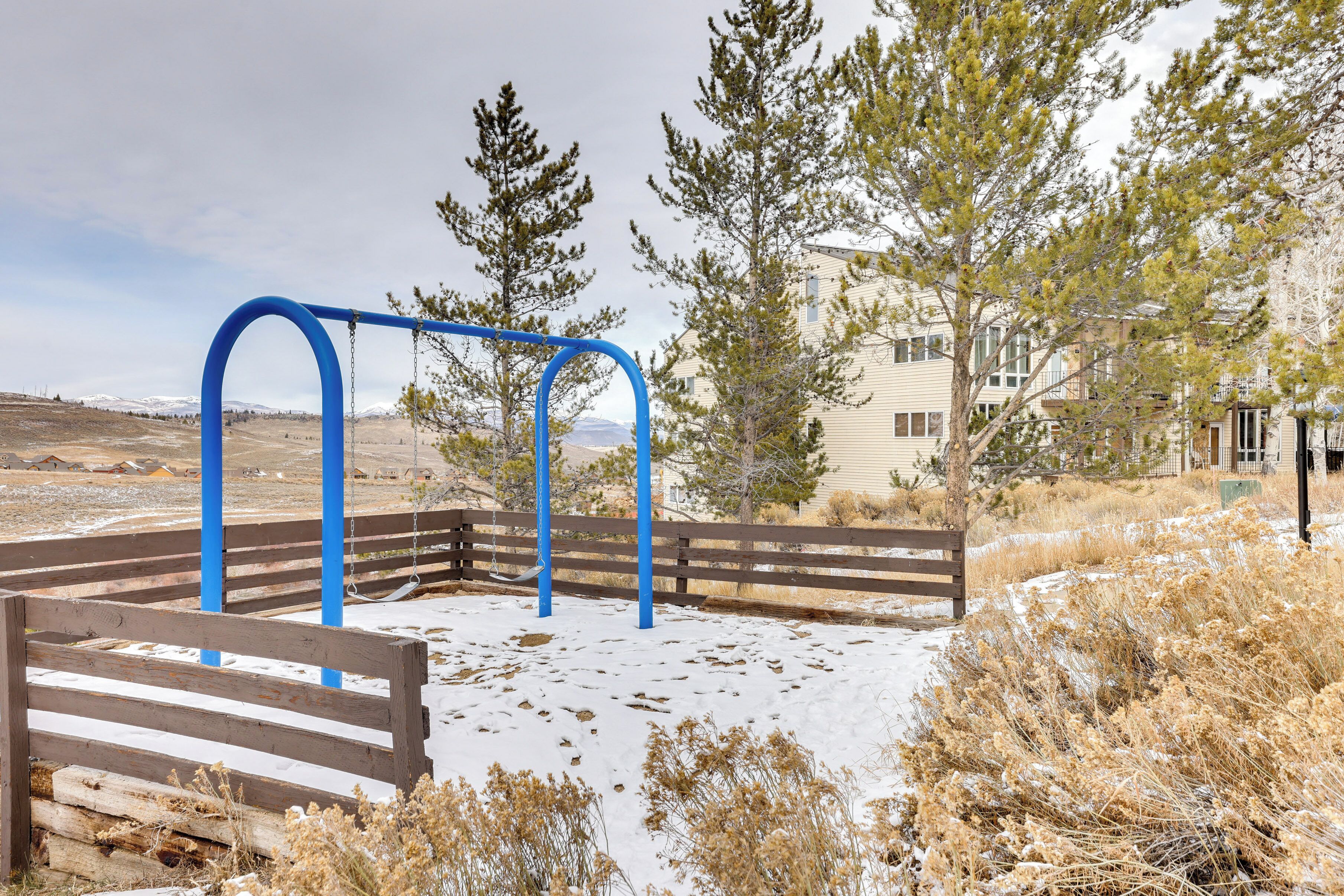 A playground with a swing set and a bench, surrounded by dry grass and trees, with a building in the background.