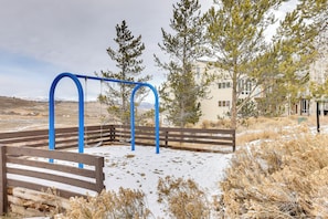 A playground with a swing set and a bench, surrounded by dry grass and trees, with a building in the background.